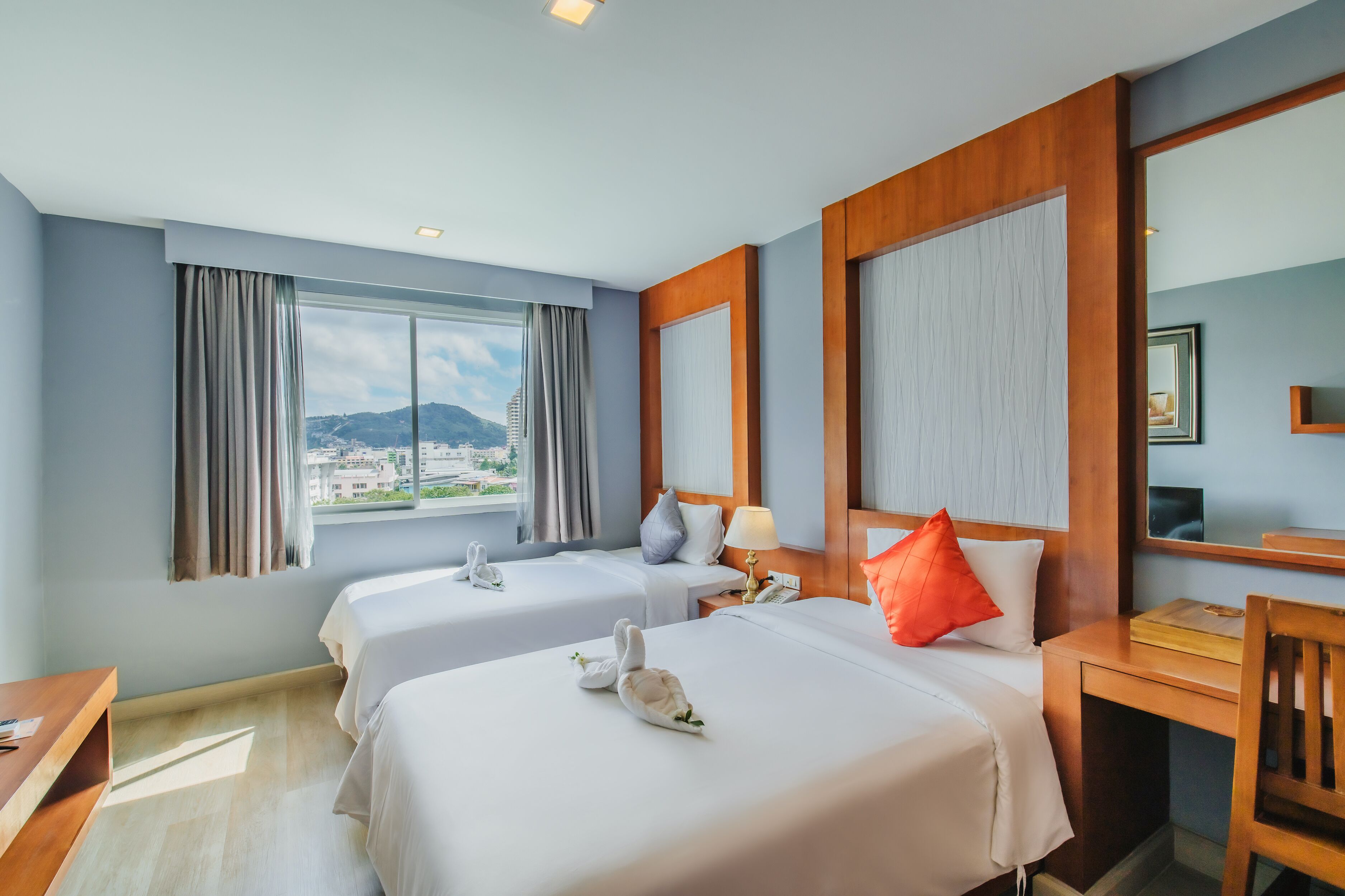 A hotel room with two beds, a desk, a chair, a window with curtains, and a view of a cityscape.