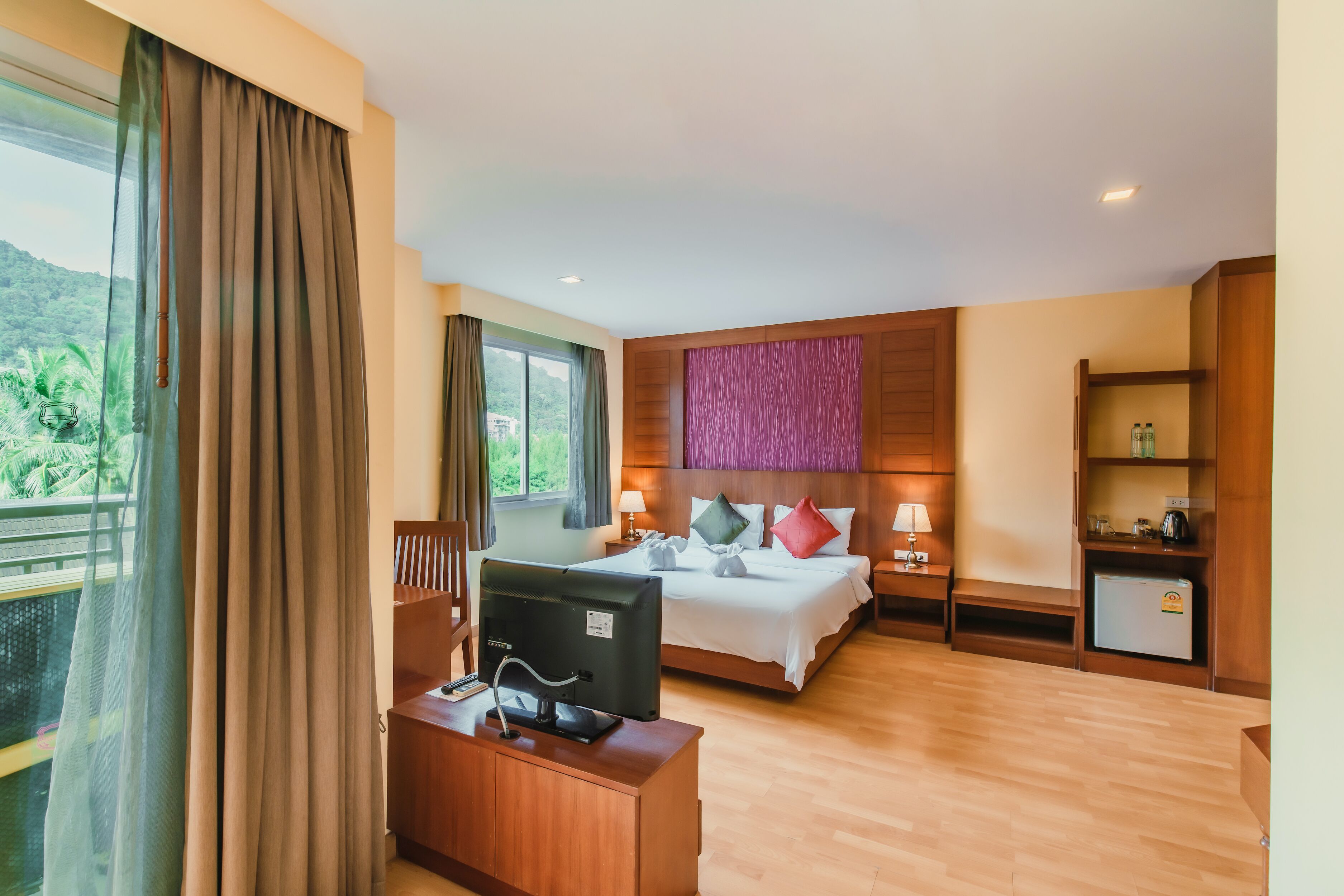 A hotel room with a bed, a desk with a computer, a small fridge, and a view of greenery outside.