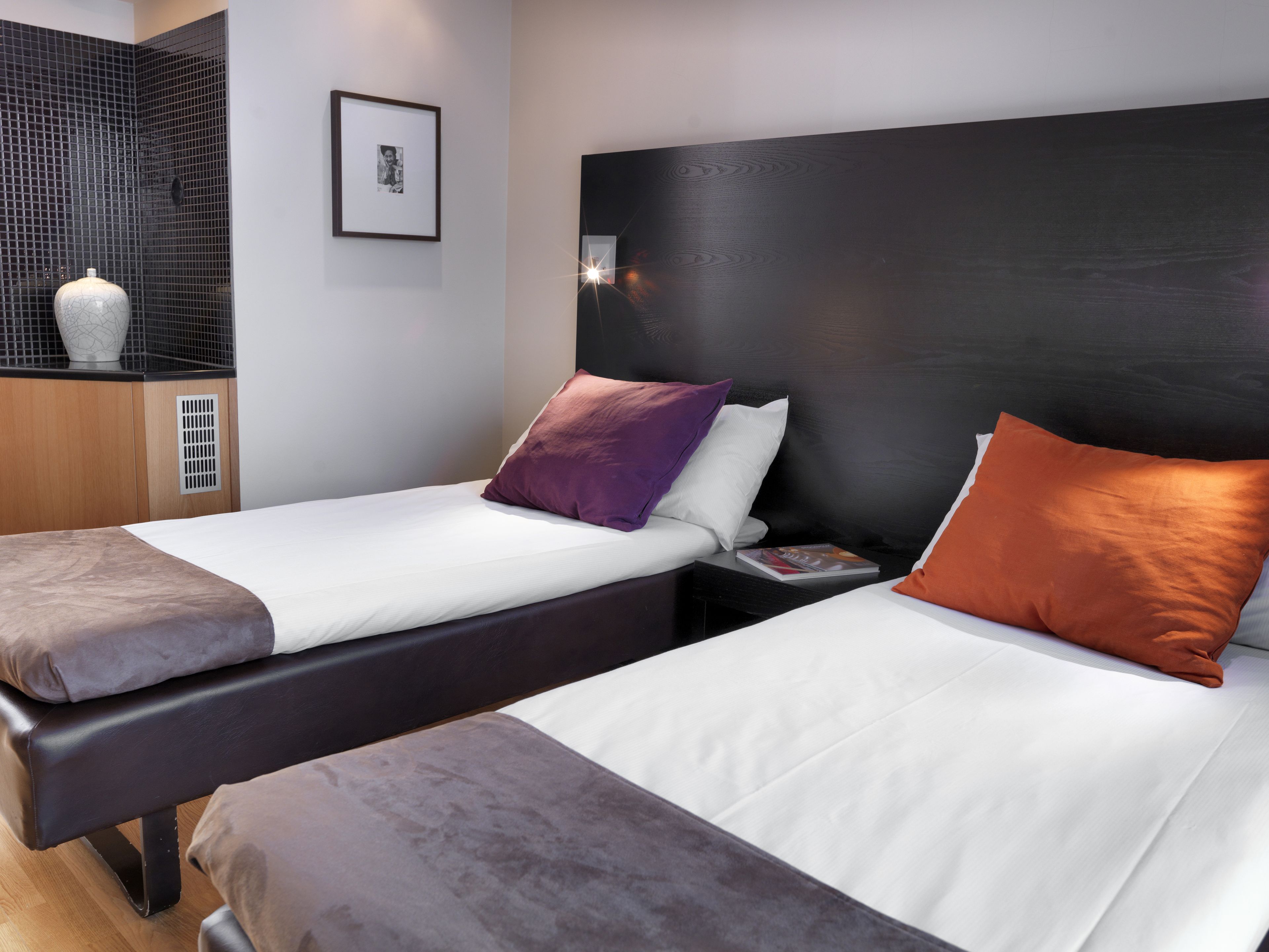 A hotel room with two beds, each with a different colored pillow, a bedside table with a lamp, and a framed picture on the wall.