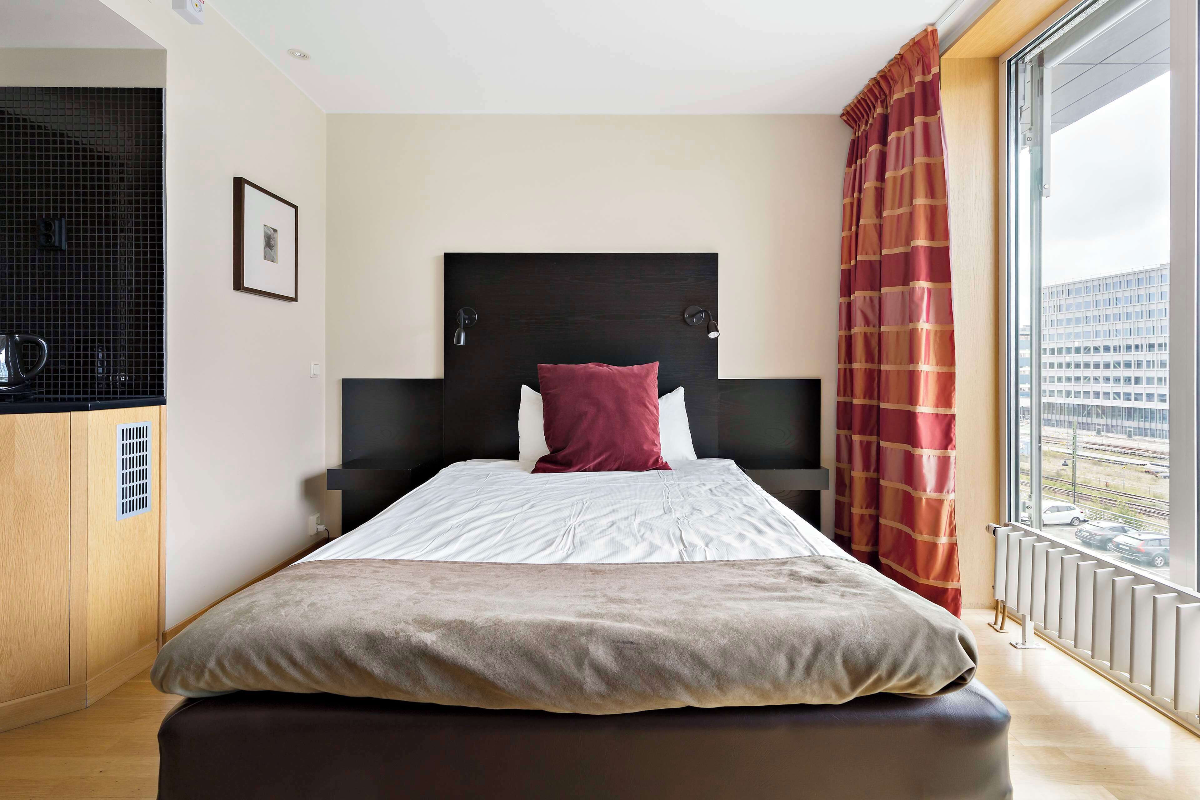 A modern hotel room with a large bed, a black headboard, a red pillow, and a view of the city through a large window.