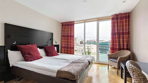 Habitación de hotel moderna con una cama grande, dos almohadones rojos, un cabecero de madera, un escritorio con silla y un ventanal con cortinas.