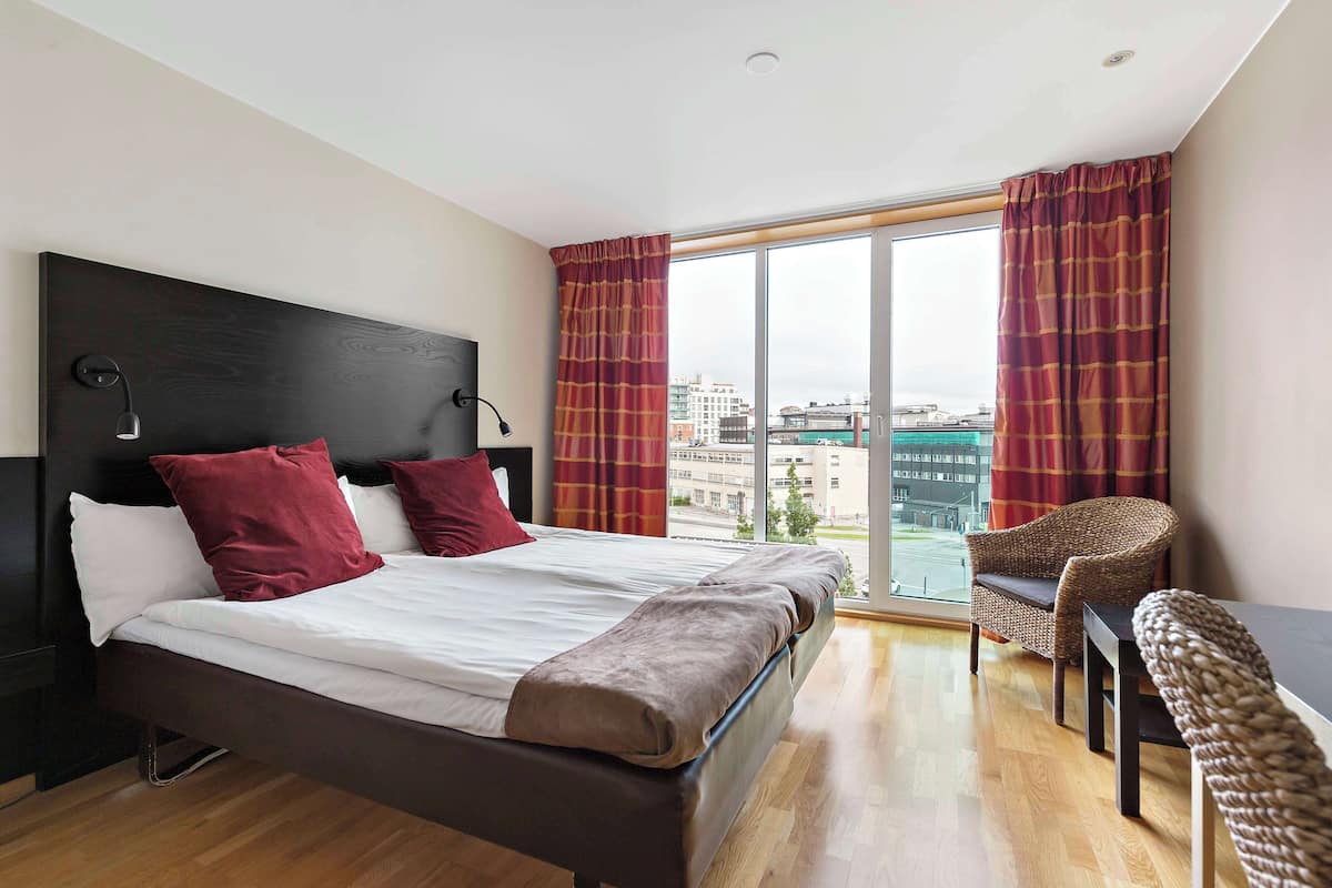 A modern hotel room with a large bed, two red pillows, a wooden headboard, a desk with a chair, and a large window with curtains.