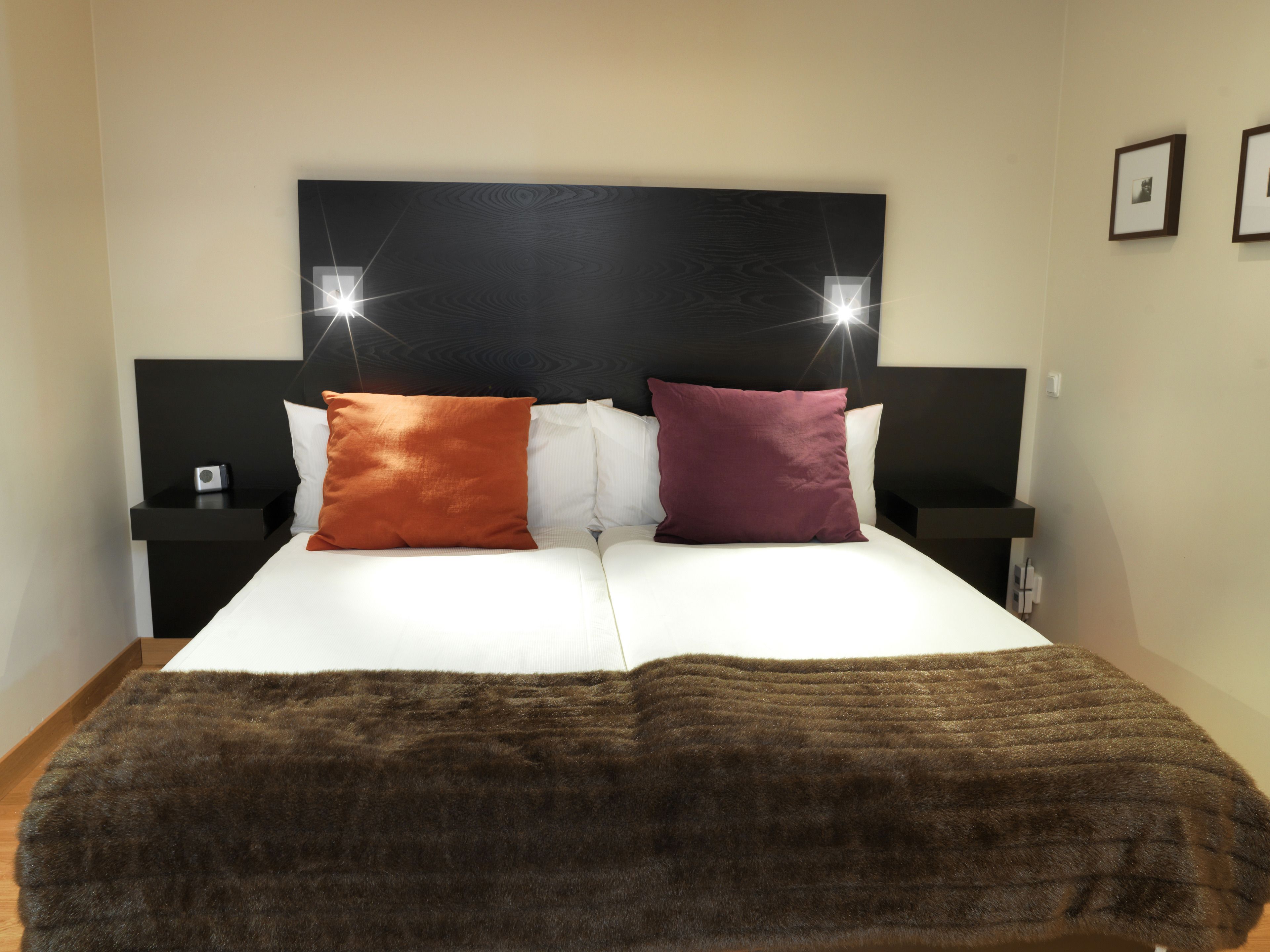 A bed with two pillows, a headboard with lights, and a bedside table with a lamp.