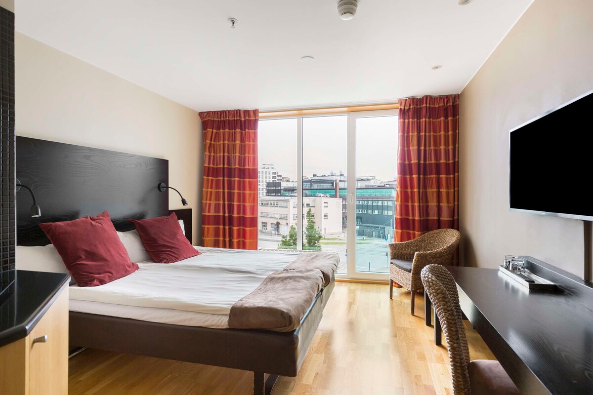 A modern hotel room with a bed, a desk, a chair, and a large window offering a city view.