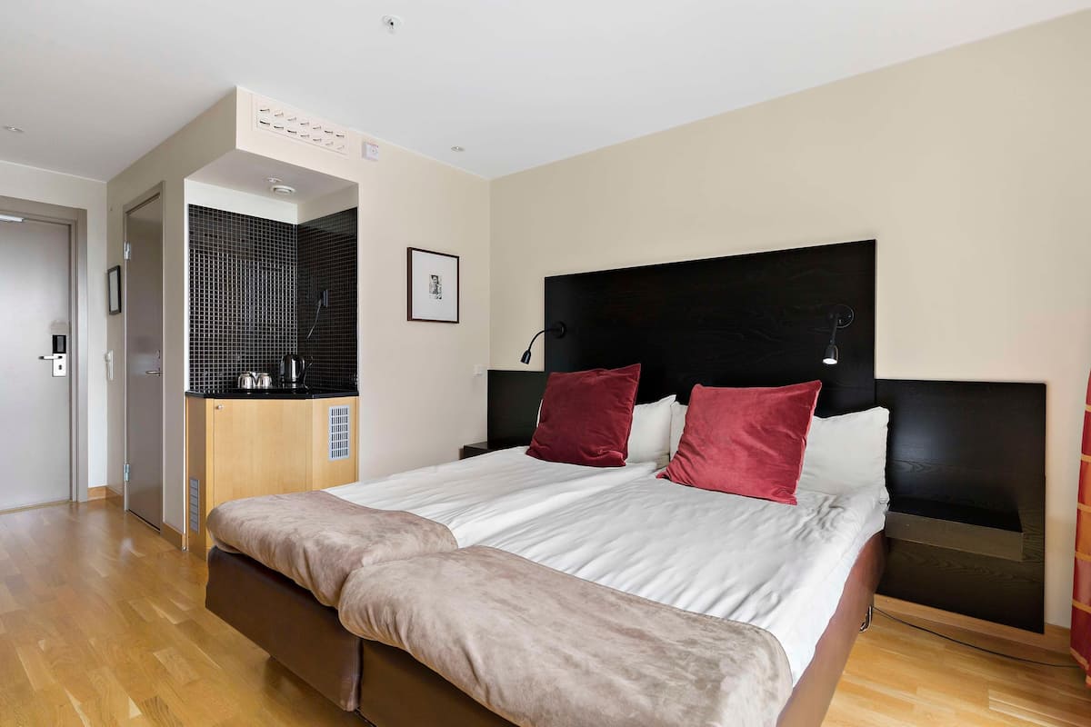 A modern hotel room with a large bed, a small kitchenette, a wooden floor, and a black headboard.
