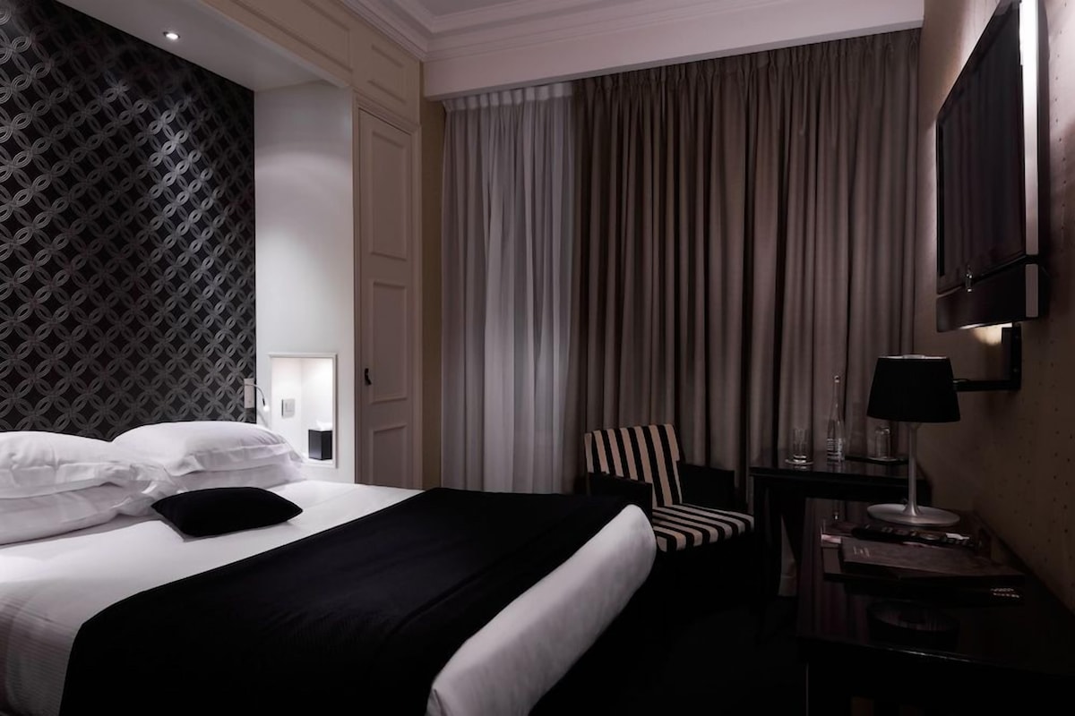 A hotel room with a bed, a chair, a desk with a lamp, and a TV mounted on the wall.
