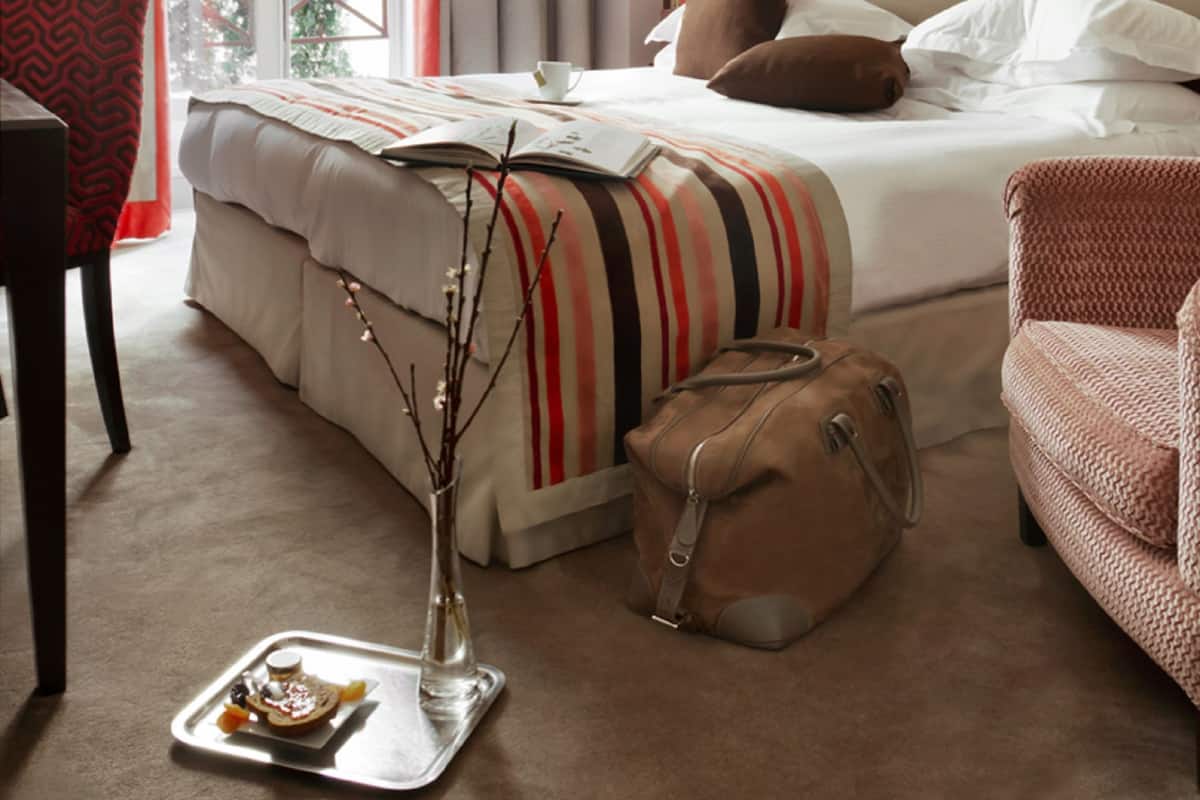 A hotel room with a bed, a chair, a suitcase, and a tray with food.