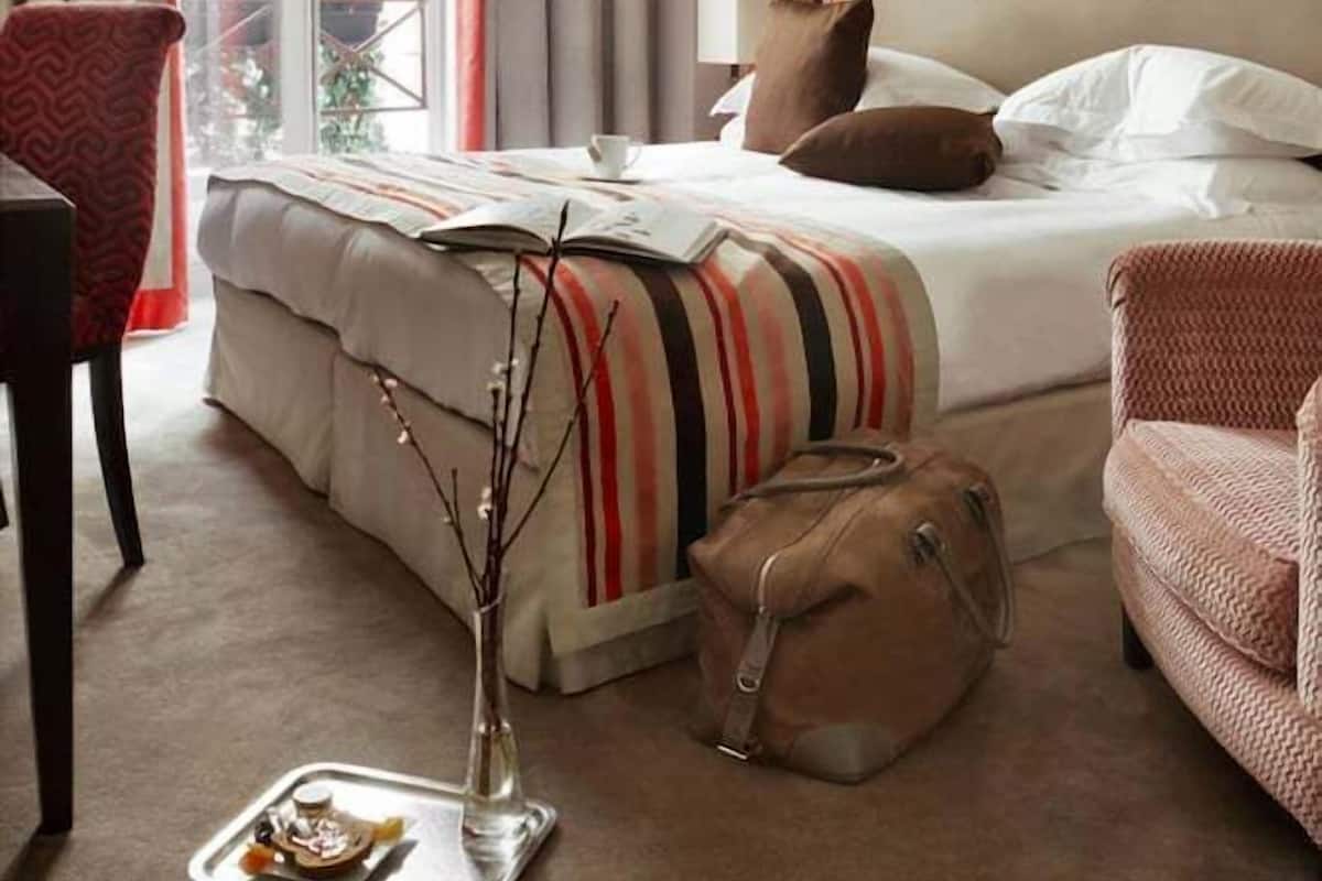 A hotel room with a bed, a chair, a suitcase, a tray with food, and a vase with branches.