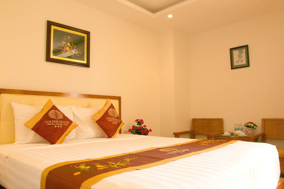 A hotel room with a large bed, two pillows with the hotel's logo, a framed picture on the wall, and a small table with a vase of flowers.