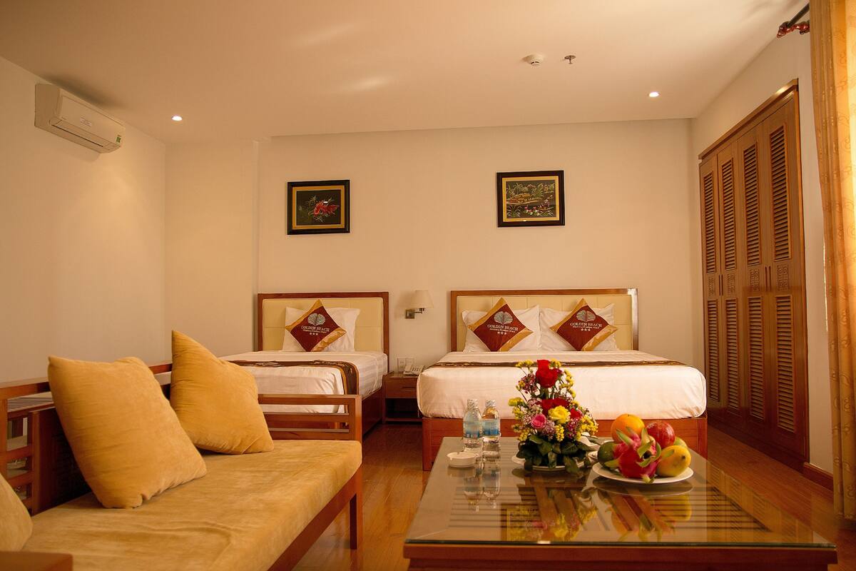 A hotel room with two beds, a sofa, a glass coffee table with fruit and flowers, and wooden wardrobes.