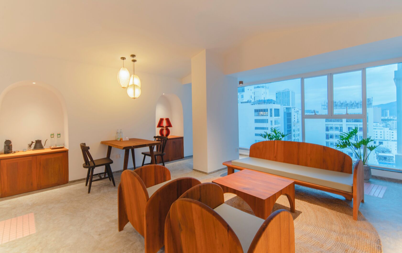 Una sala de estar moderna con un sofá de madera, una mesa de comedor con sillas, una zona de cocina y ventanas amplias que ofrecen vistas a la ciudad.