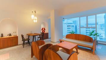 Una sala de estar moderna con un sofá de madera, una mesa de comedor con sillas, una zona de cocina y ventanas amplias que ofrecen vistas a la ciudad.