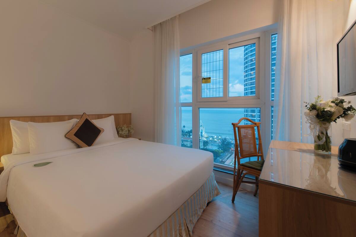 A hotel room with a large bed, a wooden headboard, a bedside table with a tablet, a chair, and a view of the city through large windows.