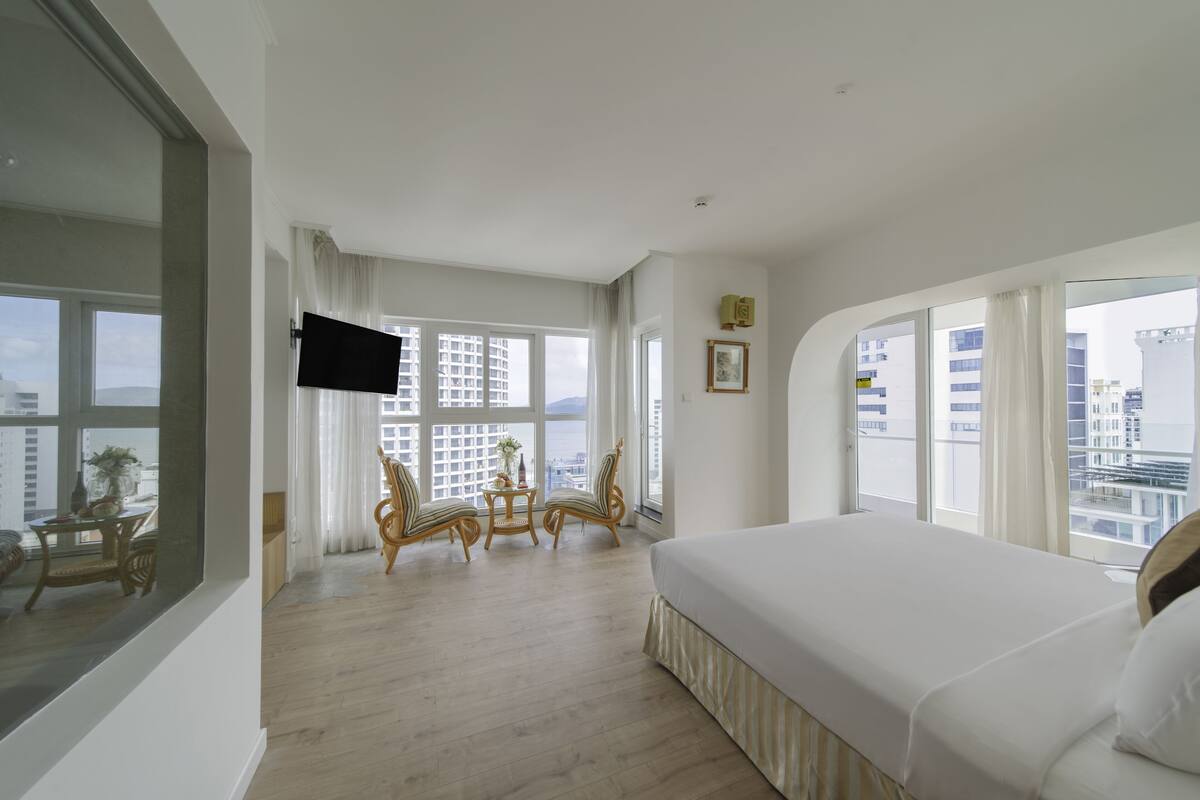 A hotel room with a large bed, a TV, a dining area with chairs, and large windows offering a city view.