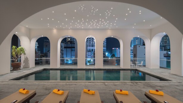 Uma área de piscina com vários espreguiçadeiras, uma piscina grande e janelas em arco com vista para a cidade.