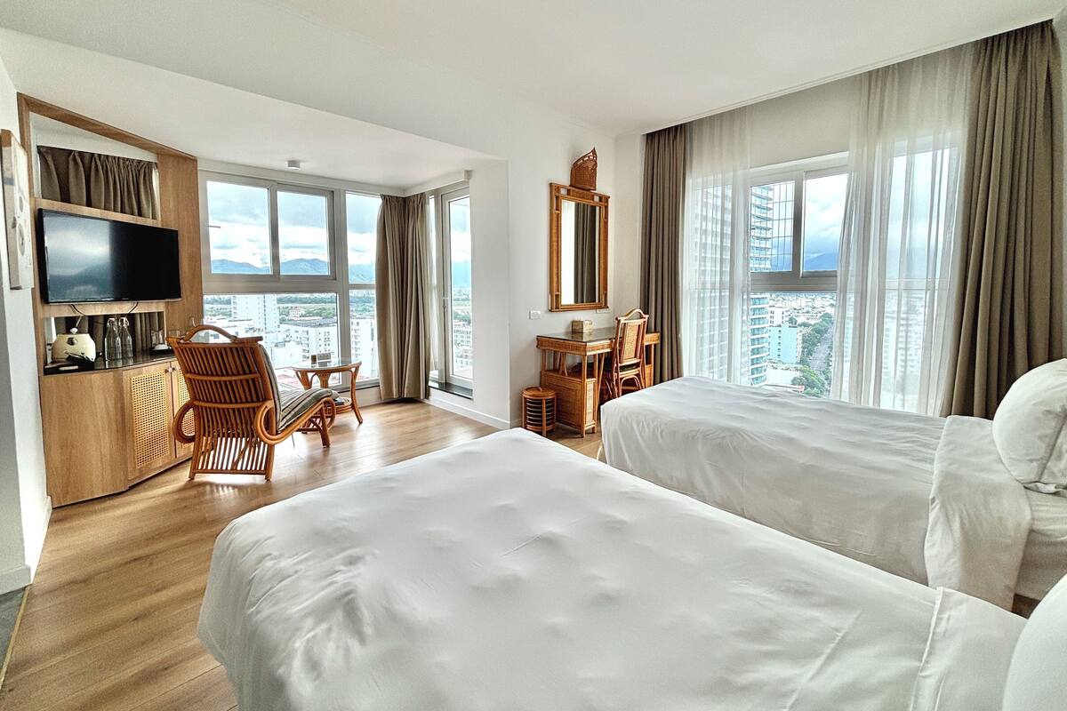 A hotel room with two beds, a desk, a chair, a TV, and large windows offering a city view.