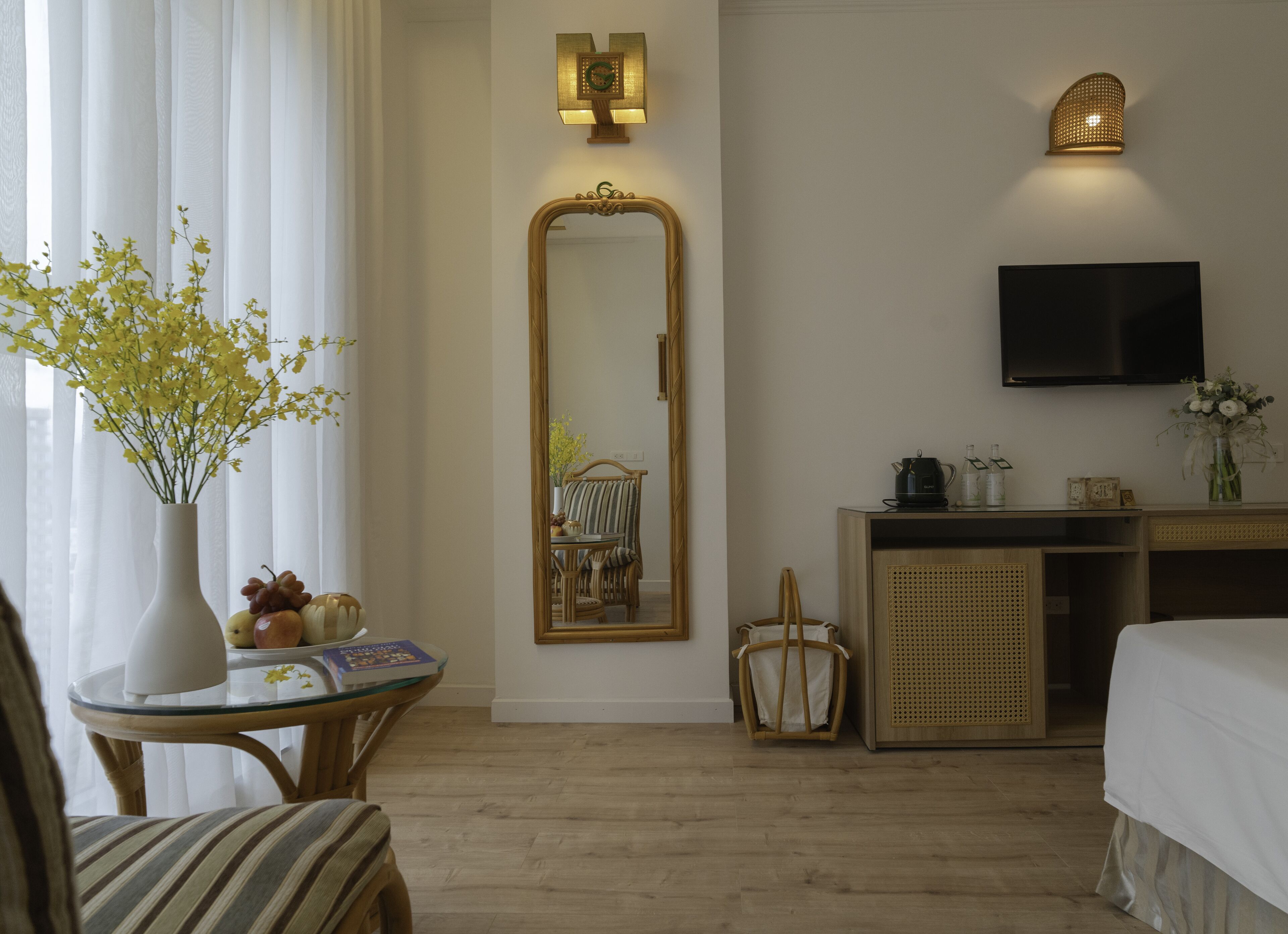 Quarto de hotel com um espelho grande, uma televisão, uma mesa de cabeceira com um vaso de flores, uma cadeira e uma cama.