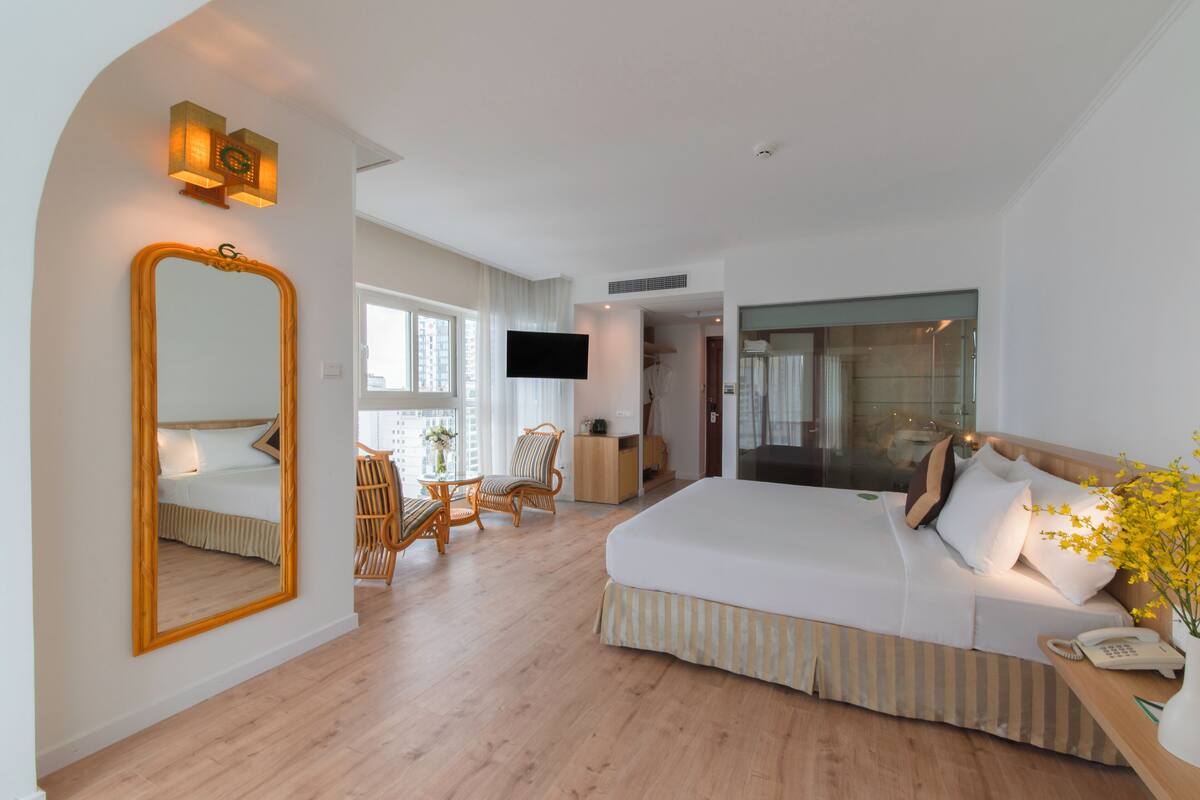 A hotel room with a large bed, a sitting area with a chair and table, a mirror, and a TV.