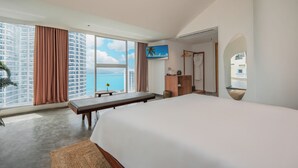 Quarto de hotel com uma cama grande, uma escrivaninha, uma cadeira, uma pequena mesa com uma flor, uma televisão e vista para o oceano e edifícios.