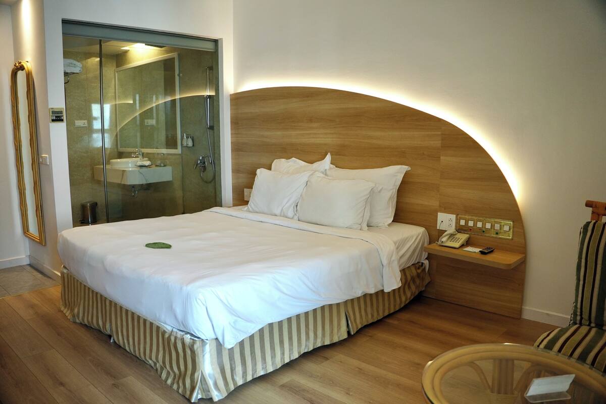 A modern hotel room with a large bed, a glass shower enclosure, a wooden headboard, a bedside table with a phone, and a striped chair.
