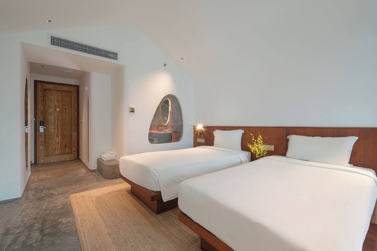 A hotel room with two beds, a wooden door, a small stool, and a wall-mounted mirror.