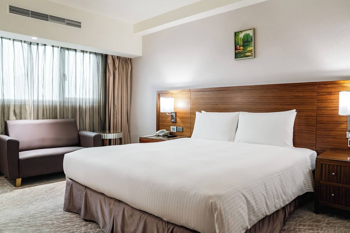Une chambre d’hôtel avec un grand lit, un canapé, une table de chevet avec une lampe, un tableau au mur et une fenêtre avec des rideaux.