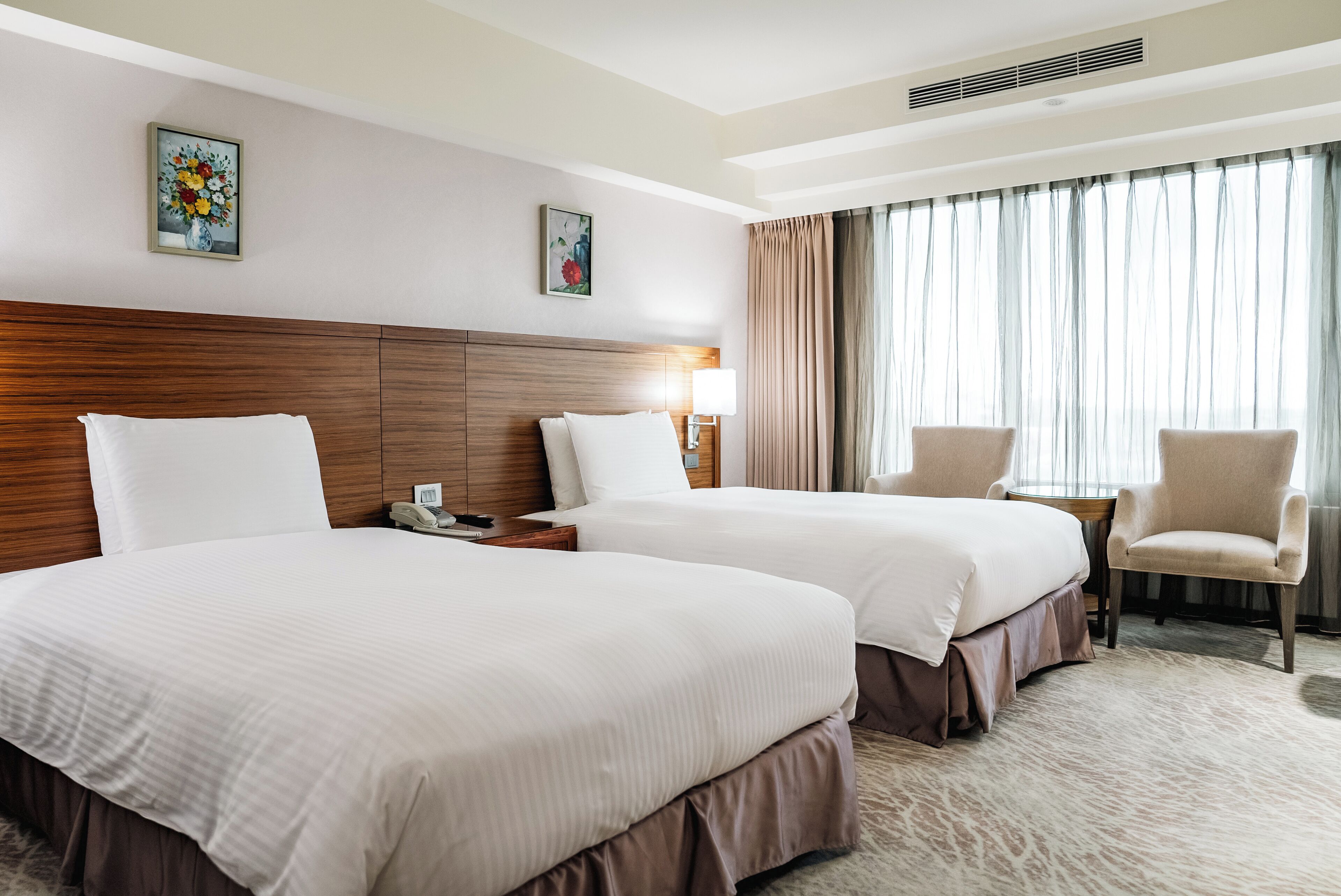 A hotel room with two beds, a wooden headboard, a small table with a lamp, a chair, and a large window with curtains.