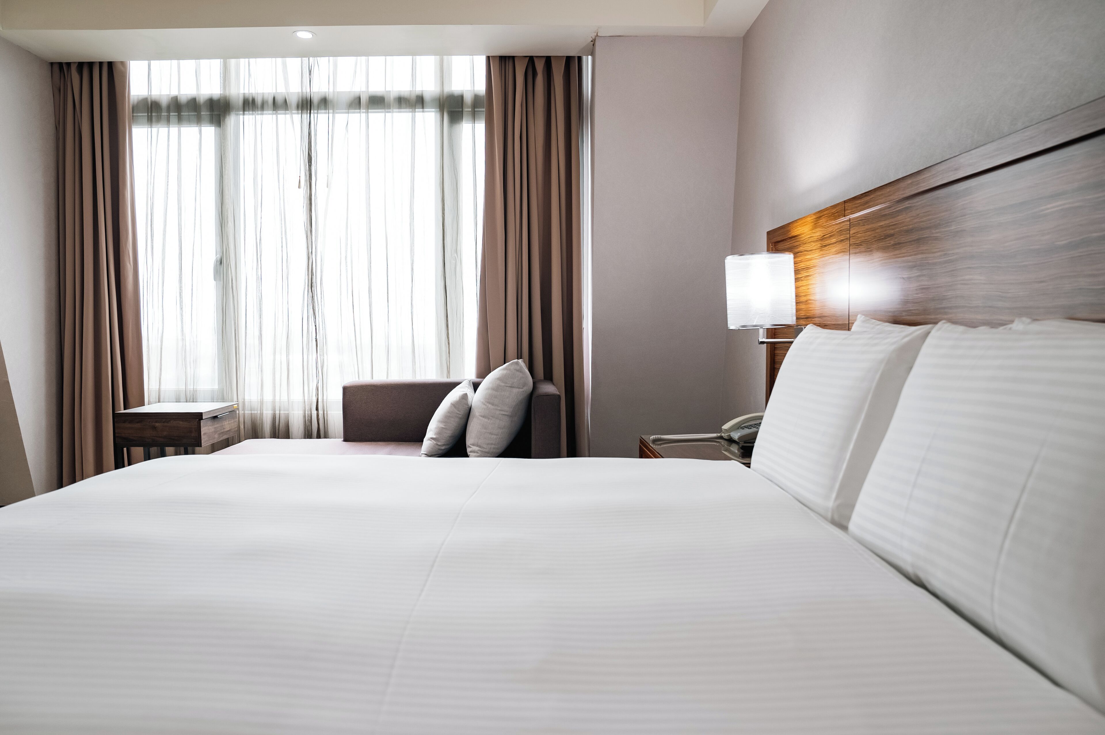 Habitación de hotel con una cama grande, una mesita de noche con lámpara, una silla y una ventana con cortinas.