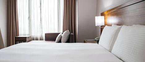 Habitación de hotel con una cama grande, una mesita de noche con lámpara, una silla y una ventana con cortinas.