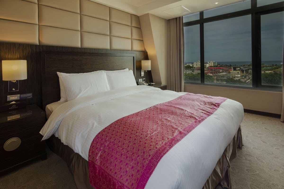 Une chambre d’hôtel avec un grand lit, une tête de lit, une table de chevet avec une lampe et une vue sur la ville grâce à de grandes fenêtres.