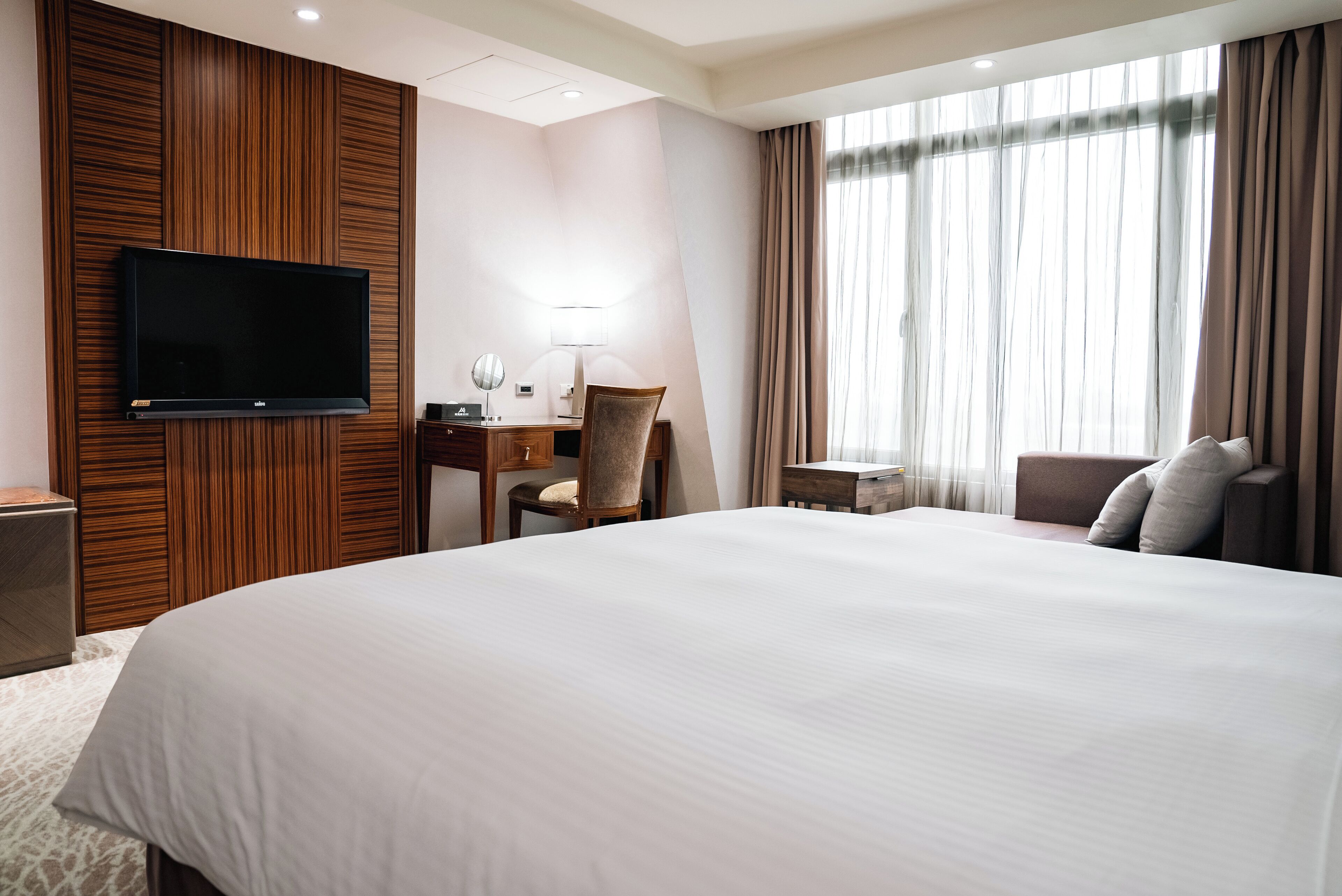 A hotel room with a large bed, a desk with a chair, a television mounted on the wall, and a window with curtains.