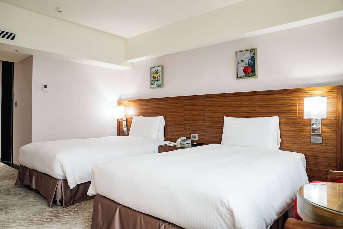 Une chambre d’hôtel avec deux lits, une tête de lit en bois, des lampes de chevet, un téléphone et une petite table avec un plateau en verre.