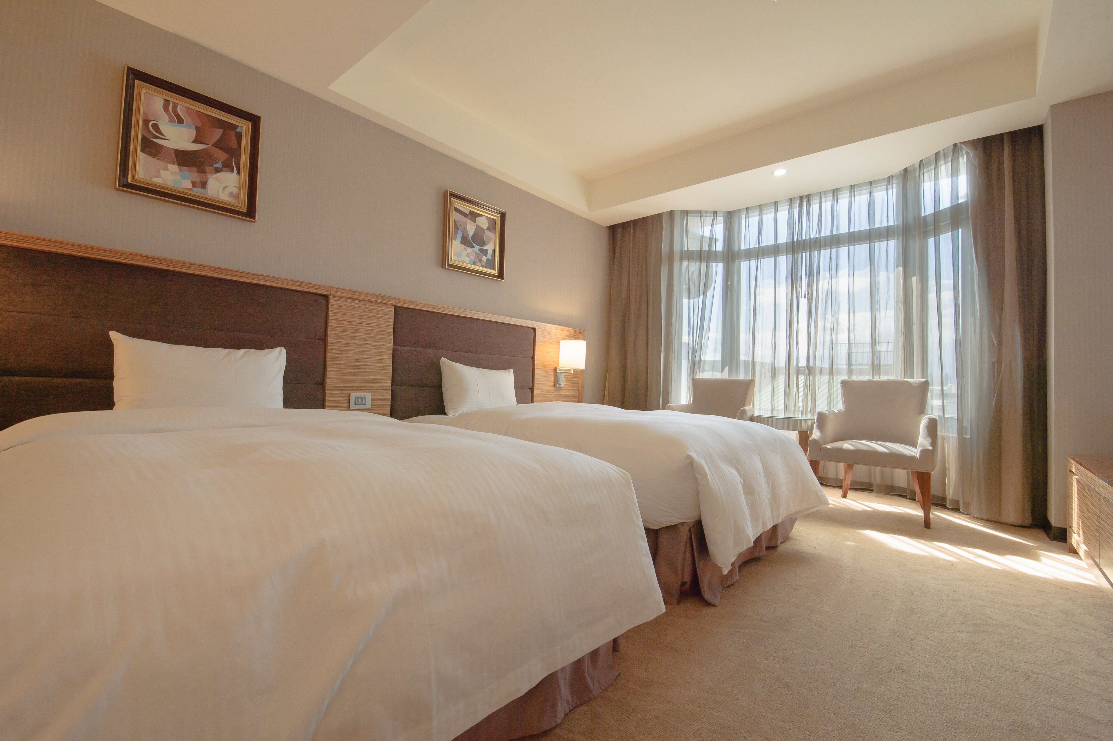 A hotel room with two beds, a chair, a desk, and large windows with curtains.
