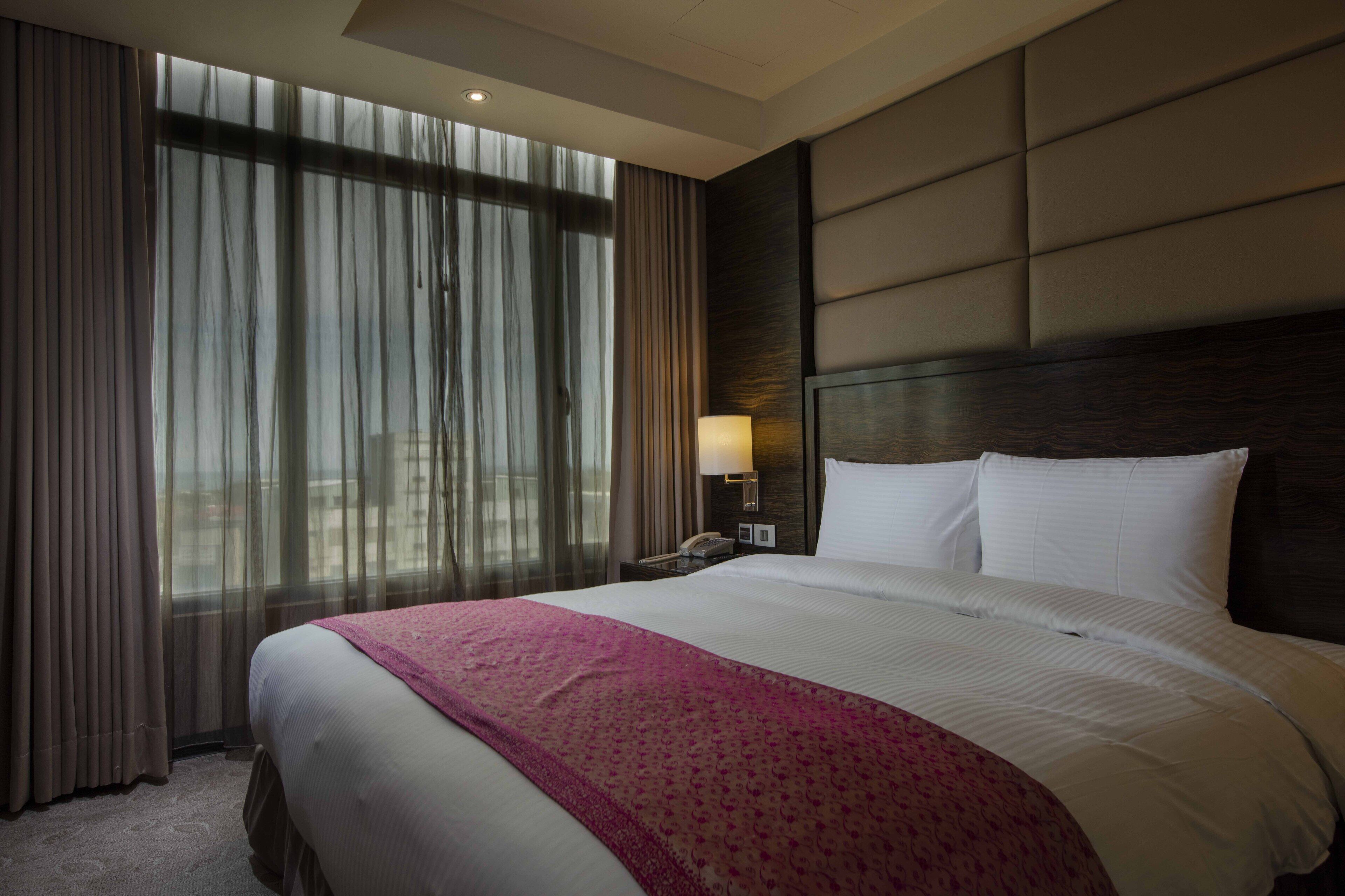 Habitación de hotel con una cama grande, una mesita de noche con lámpara y vistas a través de cortinas translúcidas.