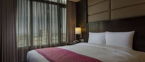 Habitación de hotel con una cama grande, una mesita de noche con lámpara y vistas a través de cortinas translúcidas.