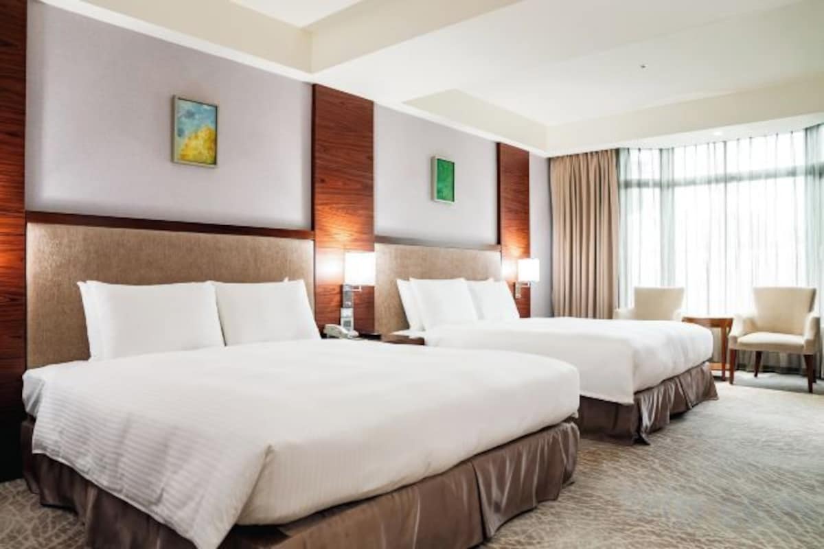 A hotel room with two beds, a desk, a chair, and a large window with curtains.