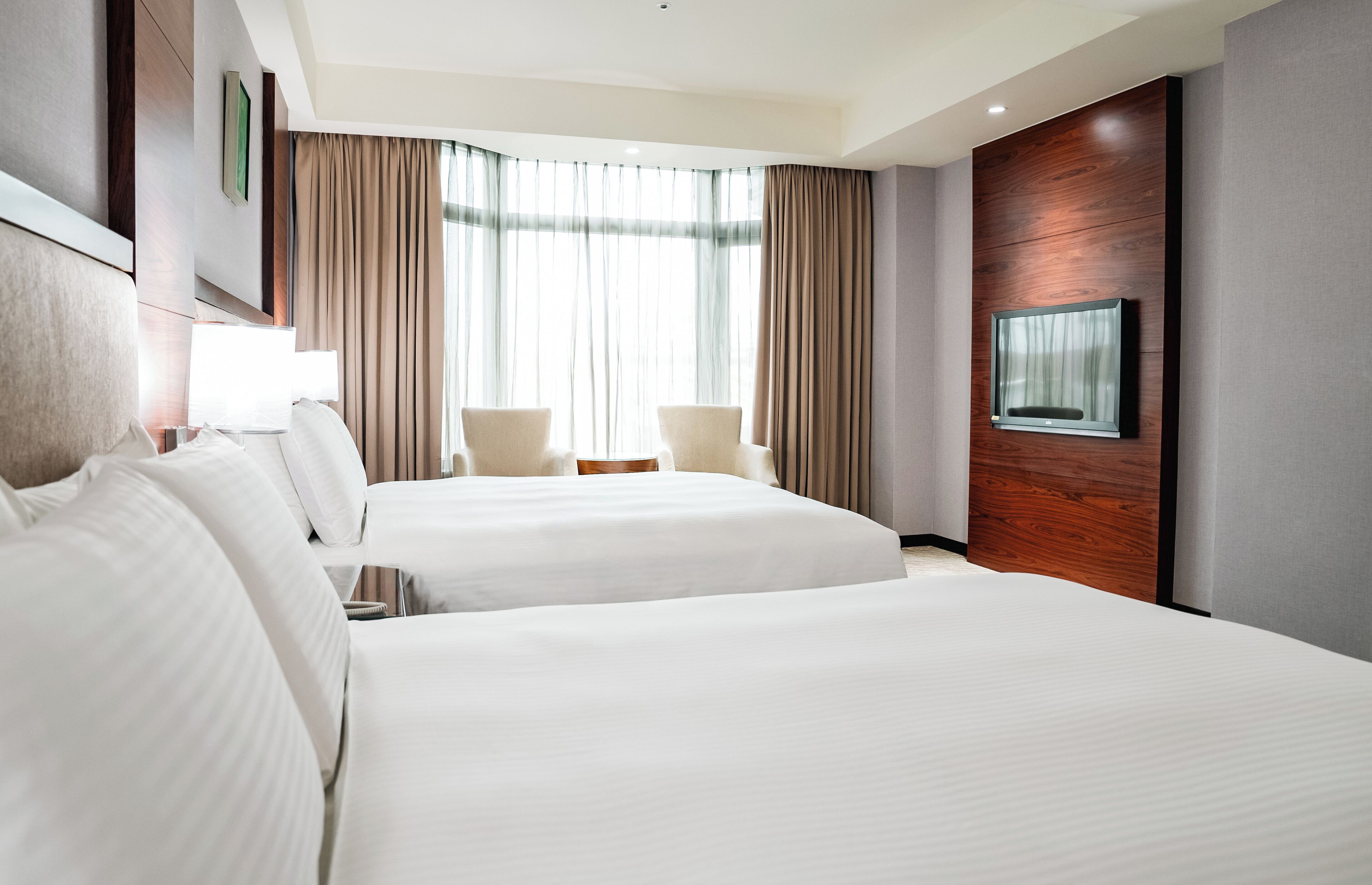 A hotel room with two beds, a TV mounted on the wall, and a large window with curtains.