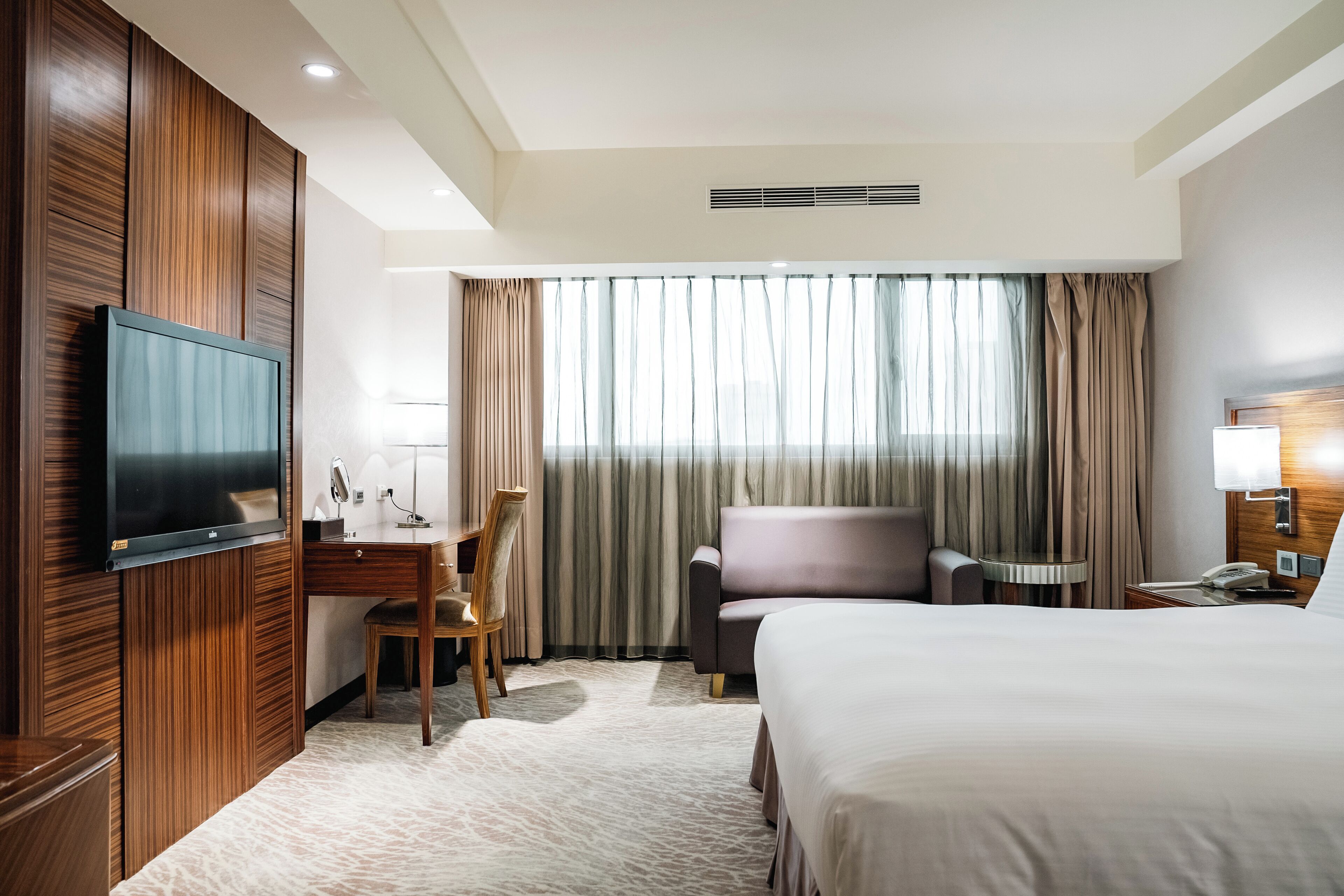 A hotel room with a large bed, a desk with a chair, a television mounted on the wall, a small table with a lamp, and a window with curtains.