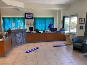 Zona de recepción de hotel con mostrador, una pantalla, un hombre de pie detrás del mostrador y una ventana con cortinas.