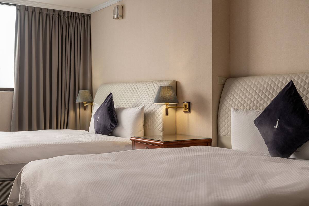 A hotel room with two beds, a nightstand with lamps, and a window with curtains.