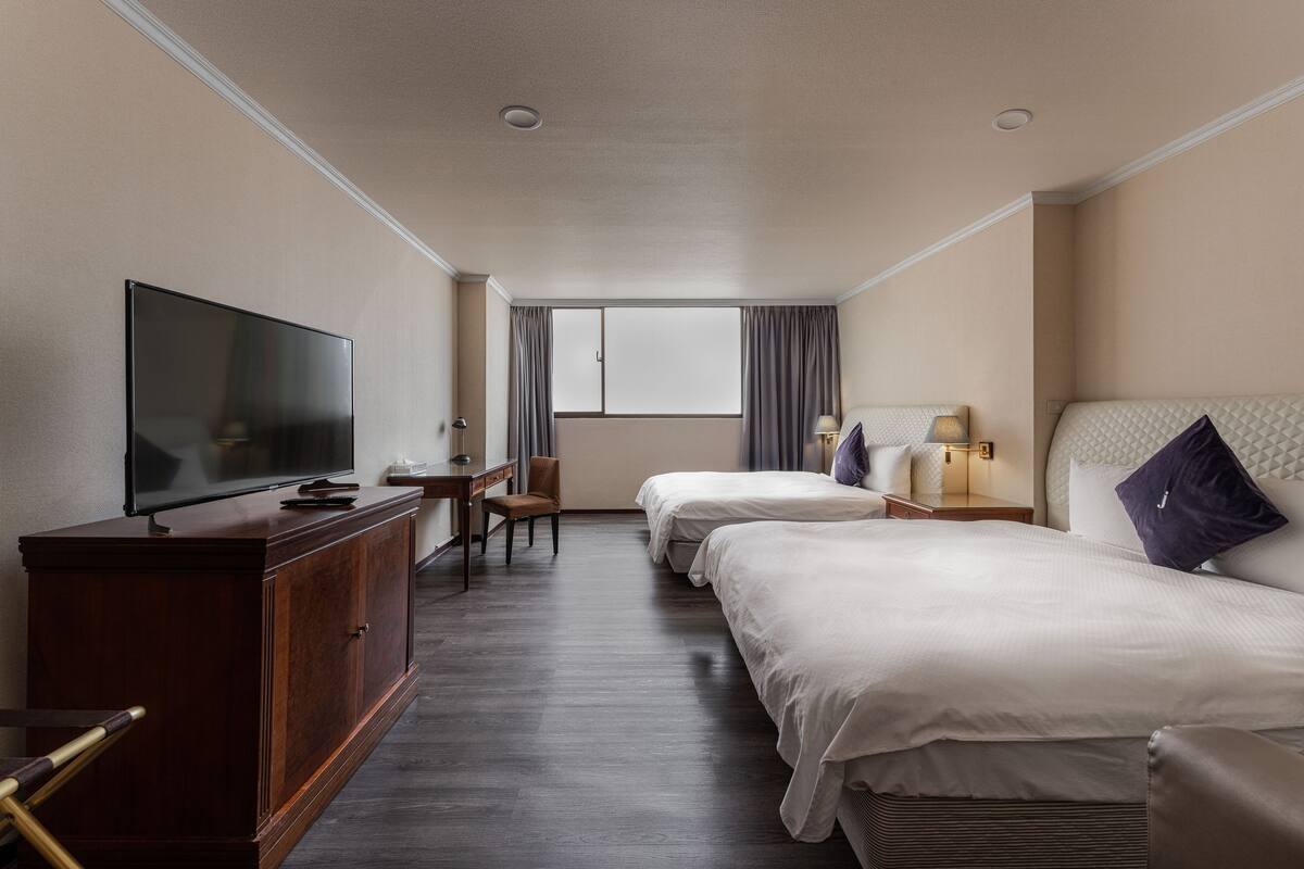 A hotel room with a TV, two beds, a desk, and a window.