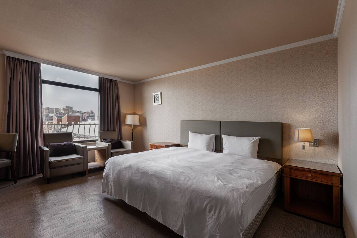 A hotel room with a large bed, a desk, a chair, and a view of the city through the window.