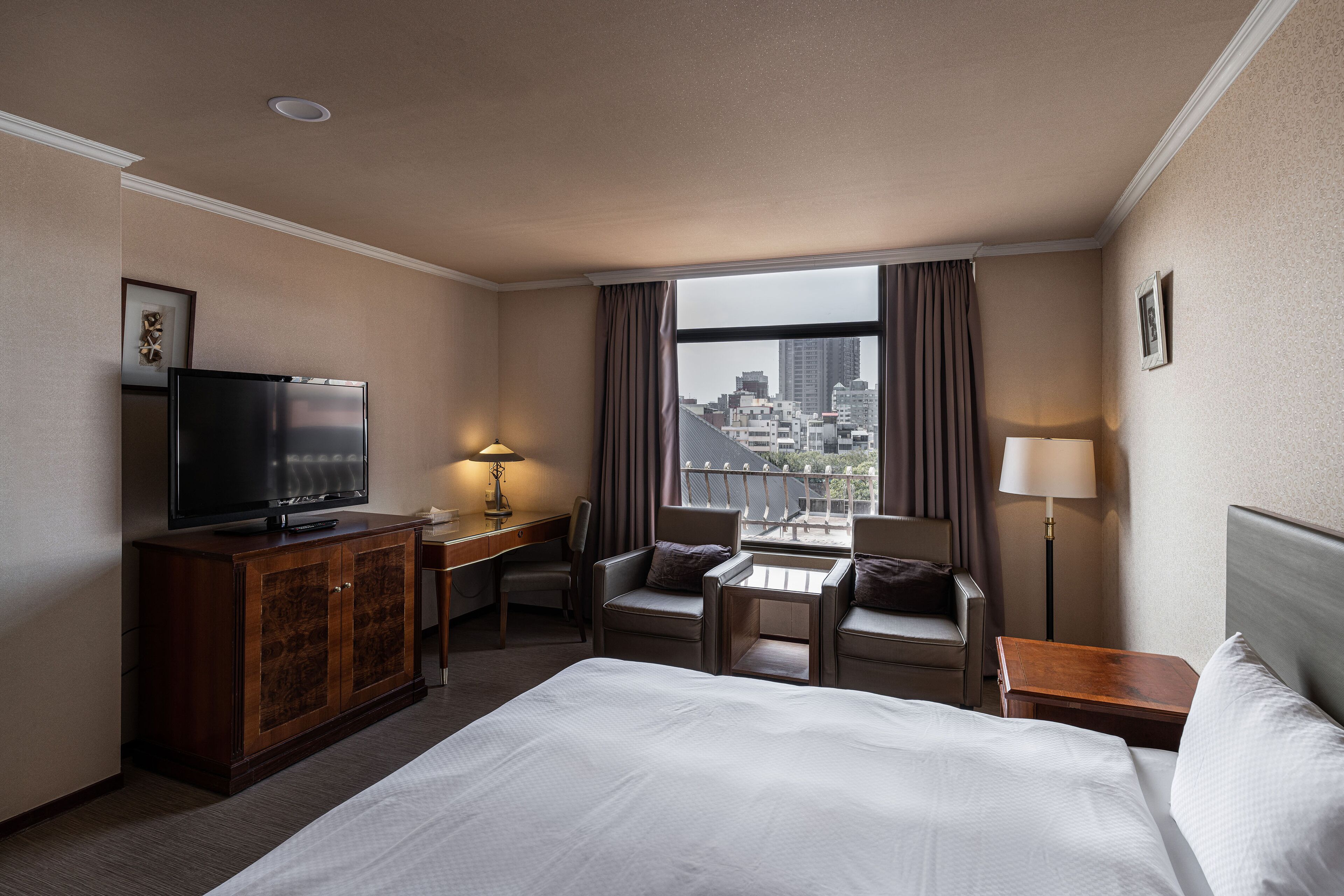 A hotel room with a bed, a TV, a desk, and a view of the city.