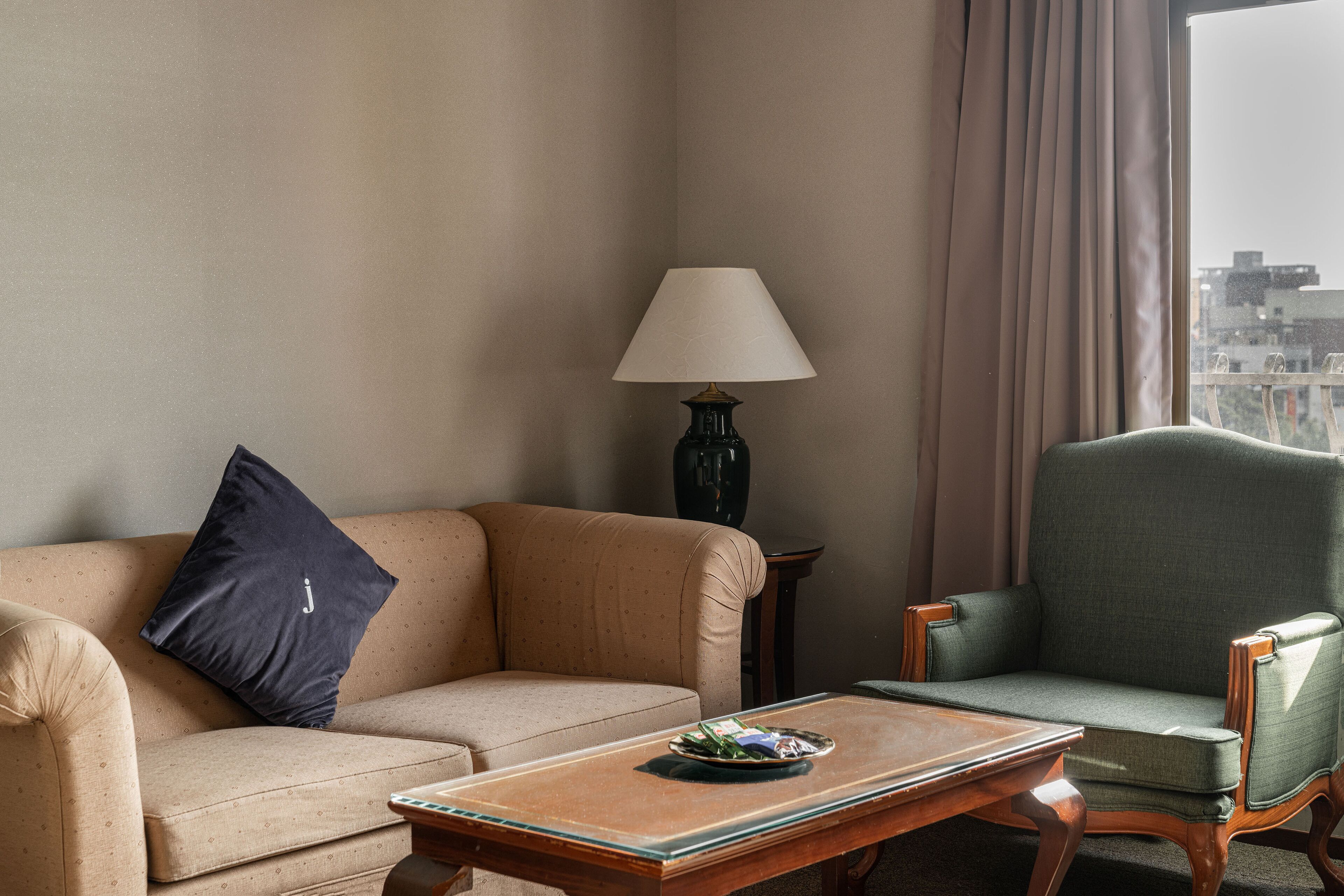 A hotel room with a beige sofa, a green armchair, a wooden coffee table, and a lamp.