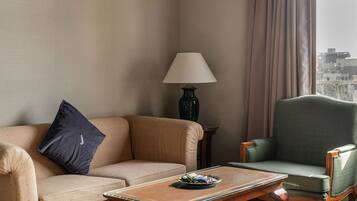 A hotel room with a beige sofa, a green armchair, a wooden coffee table, and a lamp.