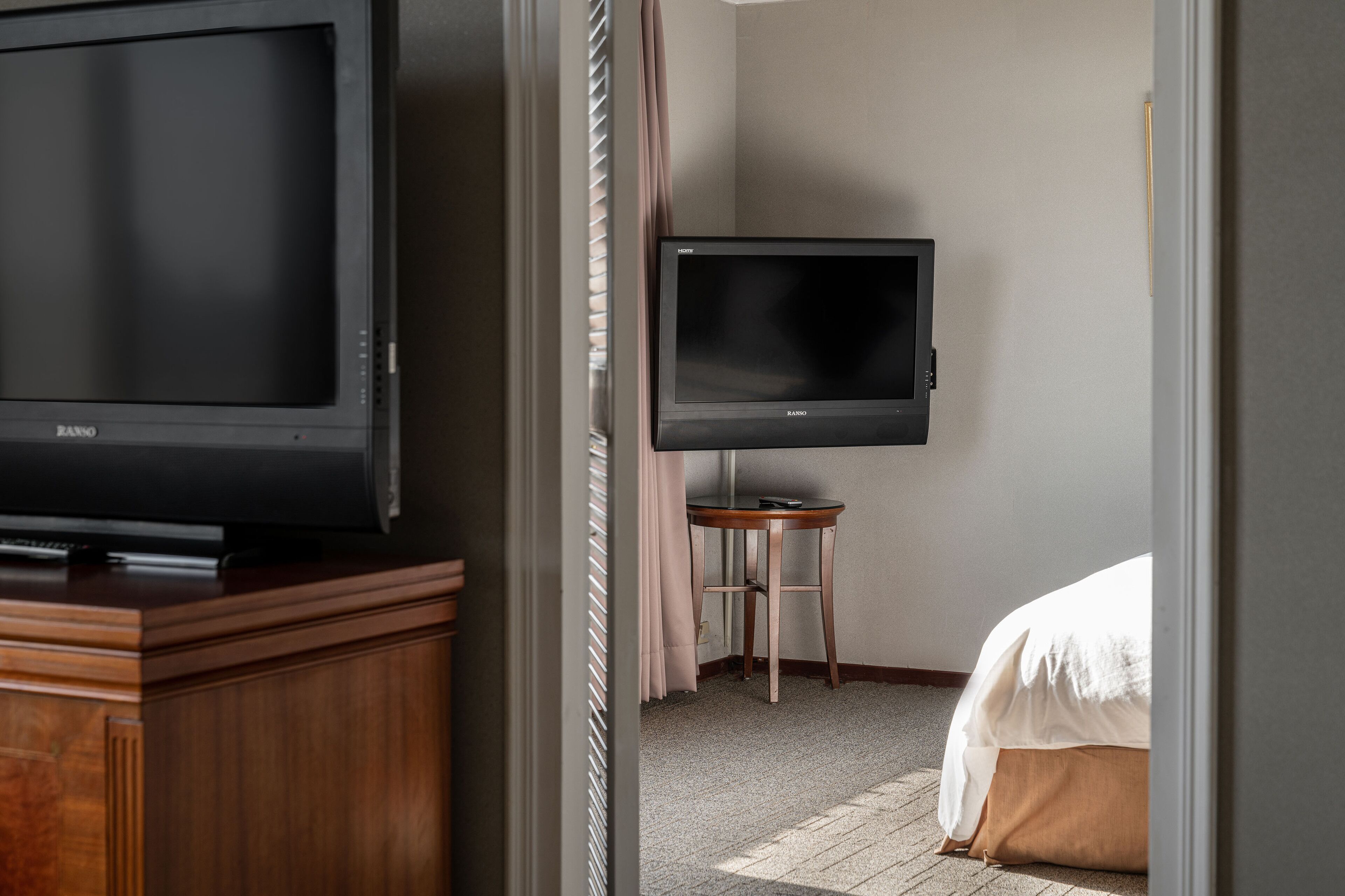 Habitación de hotel con televisión sobre un mueble de madera, una mesita pequeña y una cama con manta blanca.