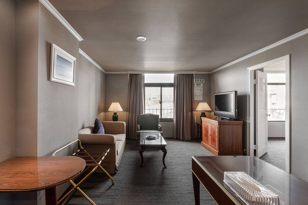 A hotel room with a sofa, armchair, dining table, TV, and a view of the city through the window.