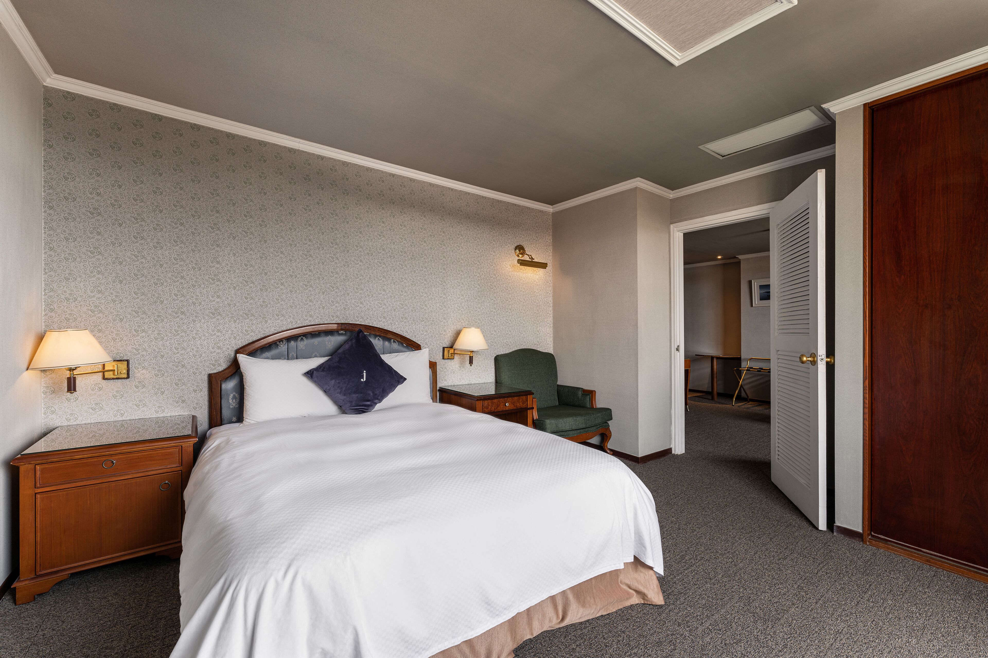 A hotel room with a large bed, two bedside tables with lamps, a green armchair, and a wooden wardrobe.