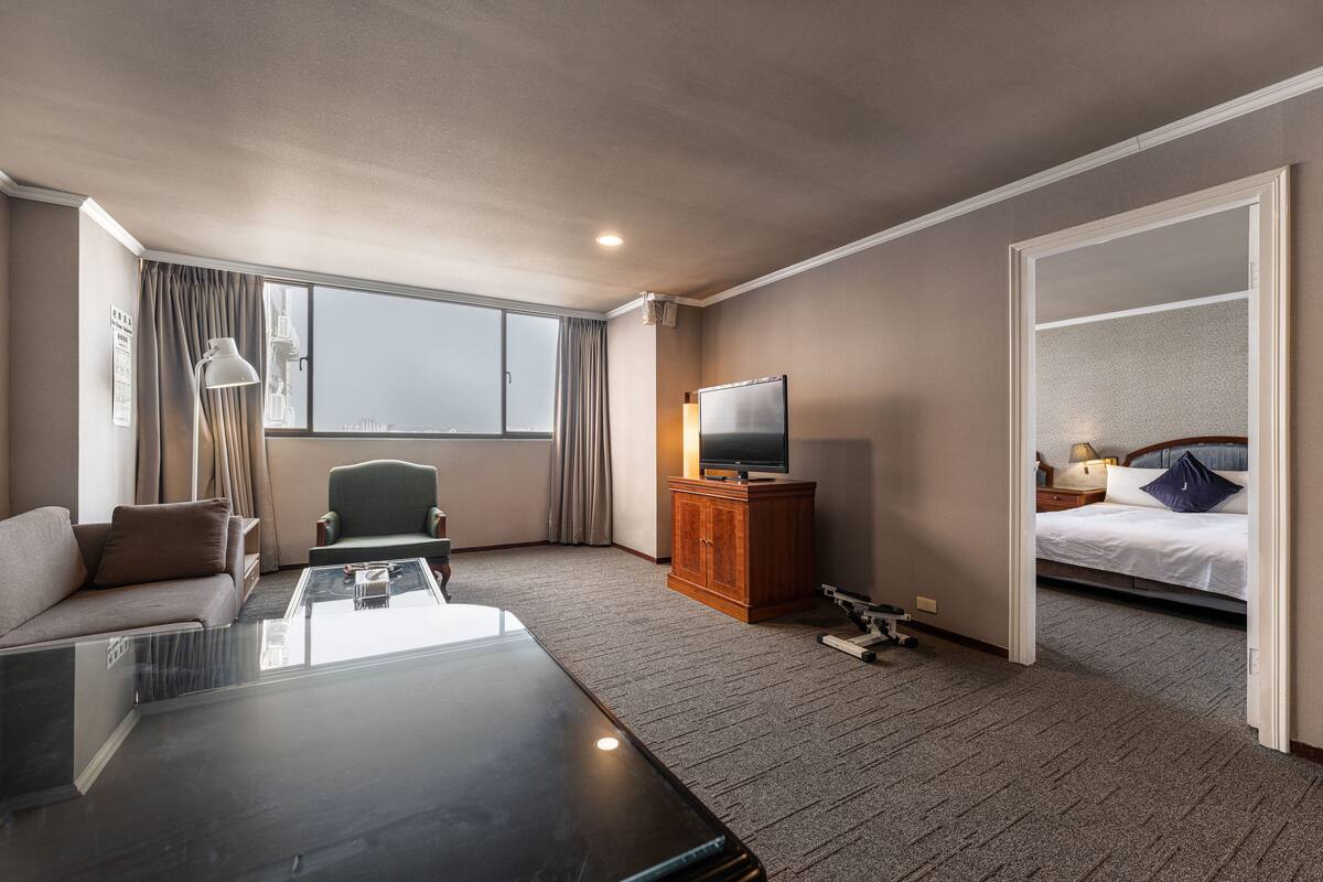 A hotel room with a sofa, a coffee table, a TV on a wooden cabinet, a bed with a pillow, and a view of a cityscape through a large window.