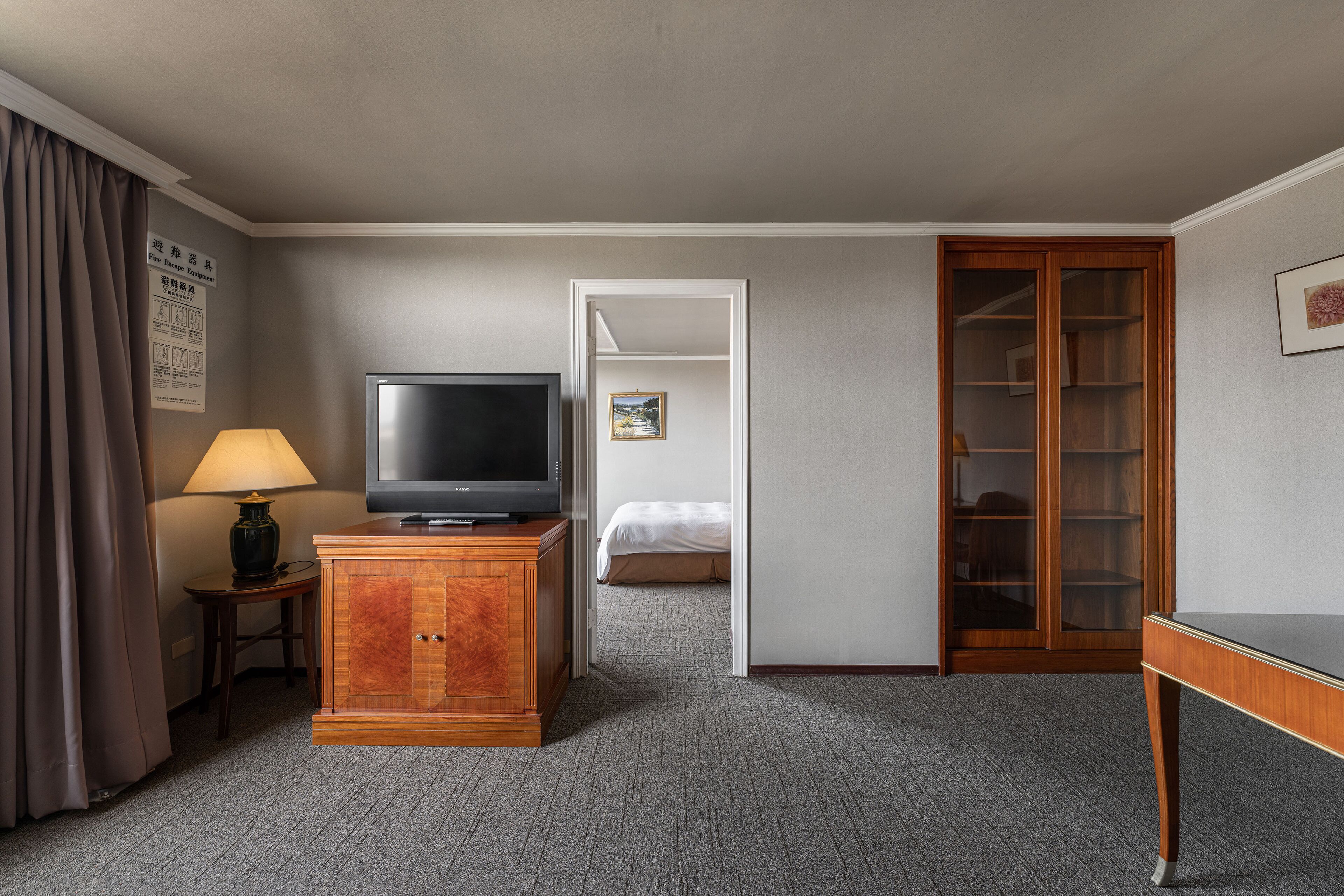 A hotel room with a television on a wooden cabinet, a lamp, a bed, and a wardrobe.